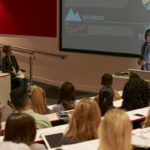 Wide shot of a university classroom in which a male professor lecture with PowerPoint slides
