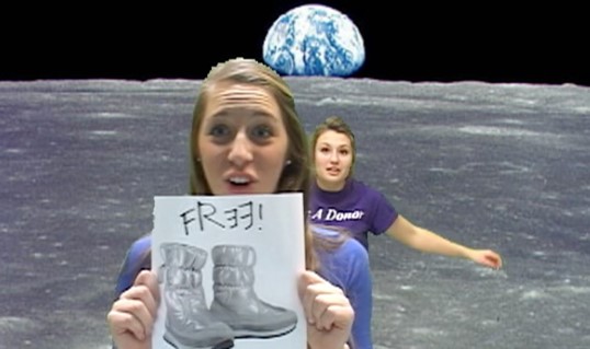 Via green screen magic, two female students stand on the moon, with the Earth visible in the background. The student closet to the camera holds a piece of paper advertising free boots.