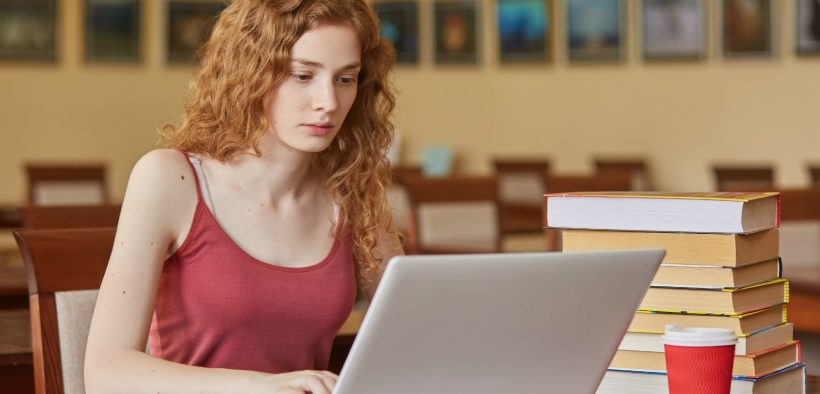 Female student blogging on a laptop next to a stack of library books