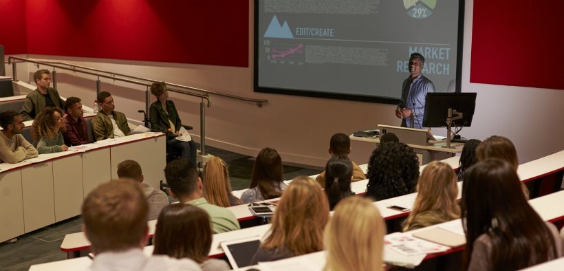 Wide shot of a university classroom in which a male professor lecture with PowerPoint slides